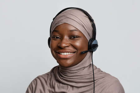 Closeup portrait of black muslim call center operator lady in hijab and headset posing over light studio background, african islamic woman help desk representative in headscarf smiling at cameraの写真素材