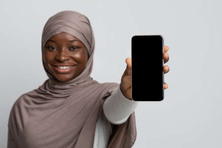 African Muslim Woman In Hijab Showing Smartphone With Black Blank Screen, Smiling Islamic Lady In Headscarf Demonstrating Modern Device With Copy Space For Your App Or Website Design, Mockup Imageの写真素材