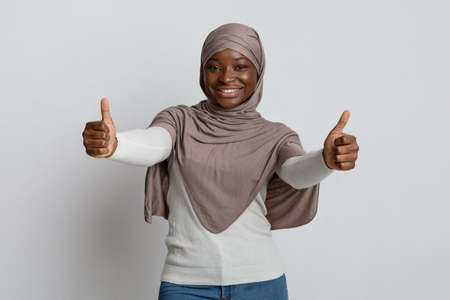 Joyful black muslim lady in headscarf showing thumbs up at camera, smiling african woman in hijab recommending great offer, gesturing sign of approval, standing over light studio backgroundの写真素材