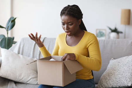 Wrong Parcel. Shocked frustrated black woman unpacking box, suffering from shipping mistake,...
