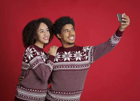Guy and lady in identical New Year outfits happily pose for selfie. Smiling young african american couple watching in smartphone, make photo for Christmas card, isolated on red background, studio shotの写真素材
