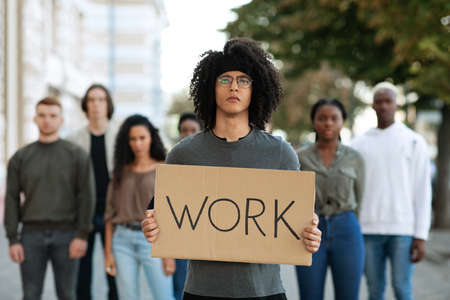 Young guy with WORK placard leading multiracial group of angry strikers. Desperate people protesting on the street, tired of lockdown, having financial troubles, anti-lockdown protests conceptの写真素材