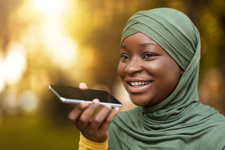 Happy Black Muslim Woman In Hijab Recording Voice Message On Smartphone, African Islamic Lady Using Virtual Assistant On Phone While Walking On City Street, Enjoying Smart Modern Technologies, Closeupの写真素材