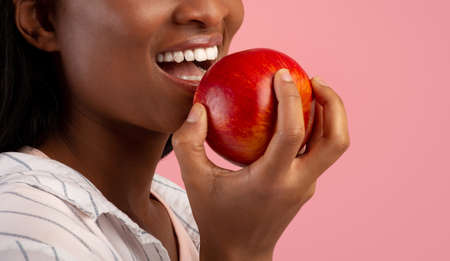 Dental care concept. Cropped view of millennial black lady with healthy teeth eating ripe red apple over pink background, copy space. Banner design. Healthy nutrition for beautiful smileの写真素材