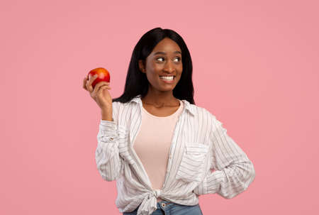 Portrait of lovely young black woman holding ripe red apple on pink studio background. Beautiful African American lady with fresh fruit, promoting healthy nutrition. Detox conceptの写真素材