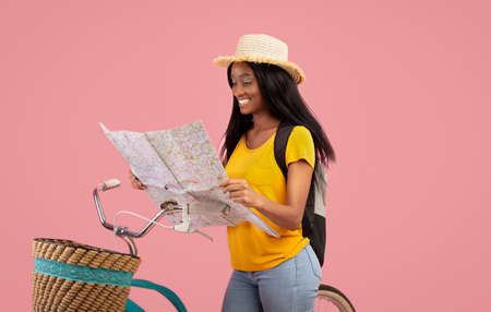 Lovely African American woman with bicycle looking at map over pink studio background. Pretty black...