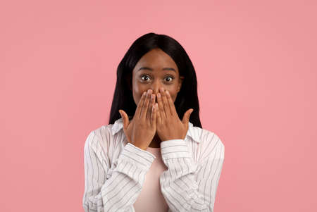 Shocked African American woman covering her mouth with hands in surprise on pink studio background. Young black lady making mistake, embarassed to hear news or gossips, cannot tell secretの写真素材