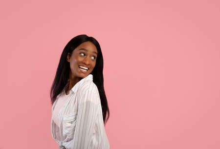 Playful black woman looking behind her shoulder at empty space on pink studio background. Portrait...