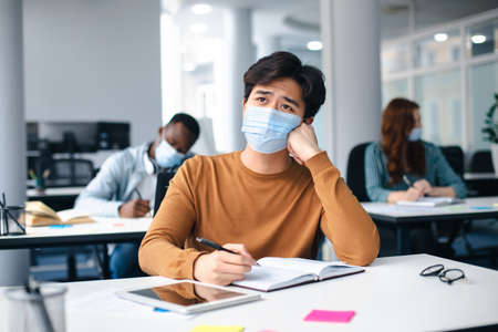 New Normal, Education And Studying Concept. Sad asian male student sitting at desk in classroom at university, wearing protective medical mask, writing in notebook and thinking, looking awayの写真素材