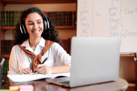 Distance Education. Smiling woman in wireless headset sitting at desk, using laptop and writing in...