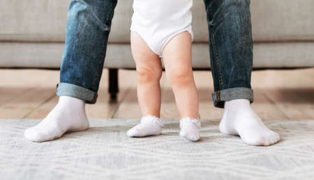 Father And Little Baby Toddler Standing Together, Loving Dad Helping His Small Daughter To Make First Step At Home. Daddys And Kids Legs. Cute Family Moments, Fatherhood Concept. Croppedの写真素材