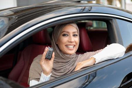 Car Owner. Joyful Muslim Woman In Hijab Smiling And Showing Automobile Keys Sitting In Her New Vehicle. Confident And Beautiful Lady Posing In Drivers Seat In Her Luxury Transportの写真素材