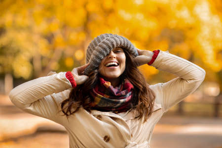 Playful young woman pulling down beanie hat on her face and laughing at park on warm autumn day....