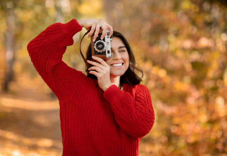 Portrait of cheerful female photographer taking photos of beautiful autumn with retro camera at autumn park. Lovely millennial lady photographing bright nature on warm fall dayの写真素材