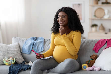 Excited pregnant black woman sitting on sofa with notebook and pen, making shopping list before...