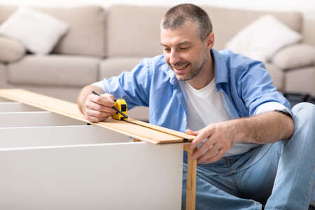 Furnishing House. Man Assembling Shelf Using Self-Assembly Kit Installing New Furniture At Home, Sitting On Floor Indoors. Handyman Services Concept. Selective Focusの写真素材