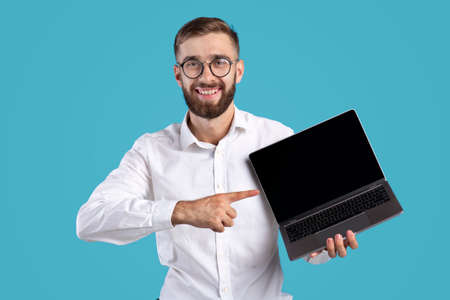 Happy millennial businessman pointing at laptop computer with mockup for website design on blue...