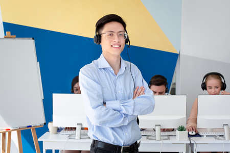 Portrait of Asian male helpdesk operator posing and smiling at call centre. Customer support agent with his diverse team working at modern open space office. Telecommunications conceptの写真素材