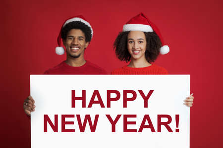 Happy New Year greetings and winter mood. Cheerful millennial african american couple in love or friends in Santa Claus hats hold billboard with inscription, isolated on red background, studio shotの写真素材