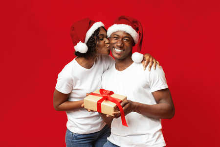 Loving black woman in Santa hat giving her happy husband New Year gift over red studio background. Emotional african american couple celebrating winter holidays together, gifts exchangingの写真素材