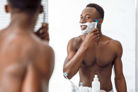Shirtless African Man Shaving Face Covered With Shave Foam Using Razor Standing Near Mirror In Bathroom At Home In The Morning. Male Facial Skincare Beauty Routine. Selective Focusの写真素材