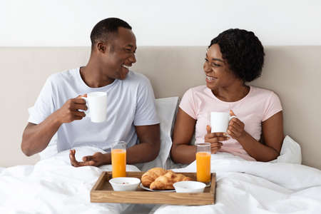 Loving african american family husband and wife having breakfast in bed at home, looking at each other and smiling. Black lovers spending morning in bed together, drinking coffee and eating croissantsの写真素材