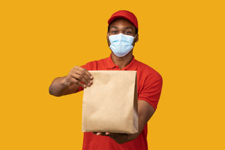 Food Delivery. Black courier in medical face mask and red cap holding and giving craft paper package from restaurant with products, isolated on yellow background. Shipment service during pandemicの写真素材