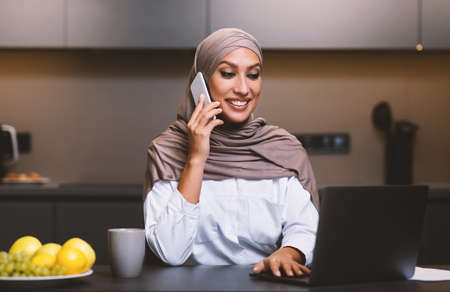Muslim Lady Talking On Phone Using Laptop In Modern Kitchen At Home, Wearing Hijab. Smiling Arab Woman Calling Restaurant Ordering Food, Browsing Internet On Computer Indoors. Selective Focusの写真素材