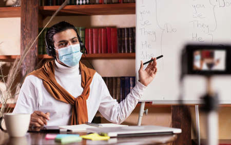 Distant Online Education. Indian Teacher Guy Making Video Lesson For Educational Blog Teaching English Grammar Rules, Pointing At Blackboard Sitting At Home, Wearing Face Mask. Selective Focusの写真素材