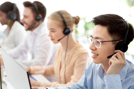 Asian hotline operator and his colleagues working at modern call centre office, copy space. Customer support agents in headsets providing services to clients, selling goods onlineの写真素材