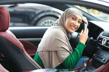 My First Car. Visiting Dealership Showroom. Beautiful muslim woman in headscarf is hugging steering wheel sitting inside her new vehicle and smiling. Young and cheerful lady enjoying purchaseの写真素材