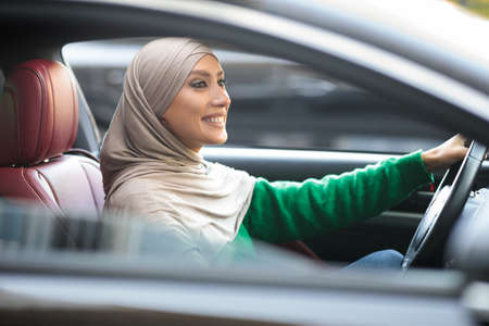 Confident Driver. Smiling Muslim Woman In Hijab Driving Modern Car In City. Young Islamic Female In...