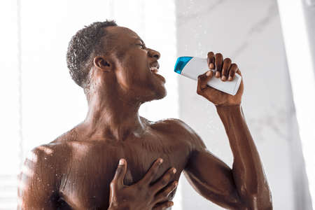Black Man Singing In Shower Holding Shampoo Bottle Like Microphone Having Fun Bathing Standing In Modern Bathroom At Home. Hygiene And Male Bodycare Beauty Routine Concept.の写真素材