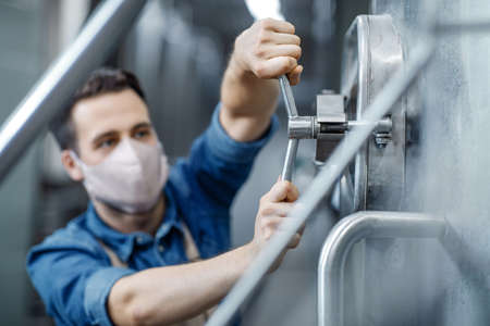 Work in plant of brewery observing process of brewing and fermentation, production of beer. Focus on equipment for drink fermentation in hands of attractive male worker in protective mask, free spaceの写真素材
