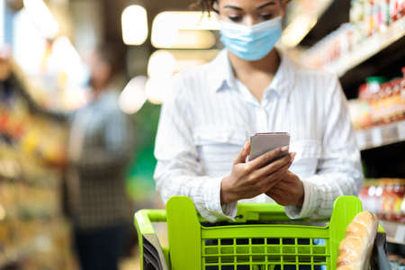African Woman In Face Mask Doing Grocery Shopping Using Smartphone, Walking With Shop Cart Buying Groceries Food Products In Supermarket Store Indoors. Shopper Mobile Application. Selective Focusの写真素材