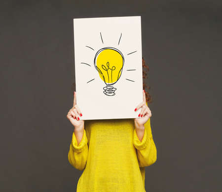 Creativity and innovations concept. Unrecognizable young woman holding poster with drawing of light bulb in front of her face, grey background. Best ideas and development strategiesの写真素材