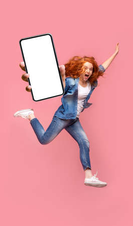 Happy young redhead woman jumping up, showing smartphone with empty screen, enjoying new application...