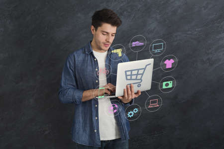 Purchasing products on internet concept. Handsome young man using laptop computer for online shopping on grey background, creative collage with icons of various household goodsの写真素材