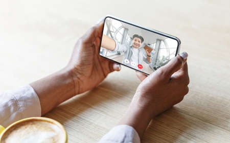 Unrecognizable black woman drinking coffee and having videocall with her caucasian boyfriend, using application on smartphone, cropped. Happy guy calling girlfriend from airport, creative imageの写真素材
