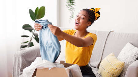 Delivery Concept. Smiling black woman holding new jeans, unboxing cardboard package, sitting on the couch in living room at home, free space. Excited lady happy with purchase and clothes shipmentの写真素材
