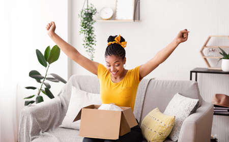 Excited african american woman received parcel, opening cardboard box at home, satisfied with great purchase, happy female model unpacking package looking inside overjoyed by postal shipping deliveryの写真素材