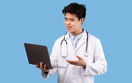 Online Consultation Service Concept. Portrait Of Confident Asian Male Doctor Using Laptop, Talking With Patient During Video Chat, Standing Isolated On Blue Studio Background, Copy Space. Telemedicineの写真素材