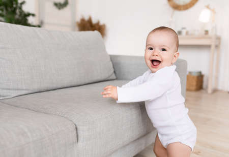 Cute Little Baby Girl Smiling To Camera Standing Near Sofa Making First Steps At Home. Happy Toddlers Portrait. Joy Of Childhood, Parenthood And Child Care Concept. Free Space For Textの写真素材