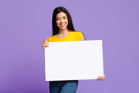 Smiling Asian Female Holding Blank White Placard In Hands, Demonstrating Copy Space For Your Text Or Design, Showing Empty Advertisement Board, Standing Over Purple Studio Backgroundの写真素材