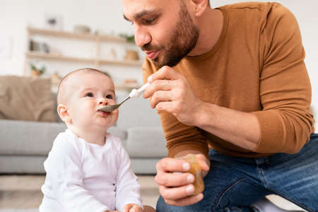 Young Father Feeding Little Baby Son With Spoon Sitting On Floor At Home. Dad Caring For Child Toddler. Paternity Leave, Fatherhood, Healthy Food And Nutrition For Newborn Babies Conceptの写真素材