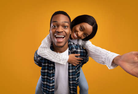 Portrait of lovers on date and have fun together. Happy young african american male holds woman on back, laughs with open mouth, lady makes selfie, isolated on yellow background, studio shotの写真素材