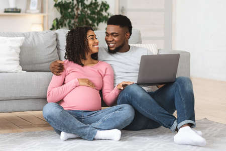 Loving pregnant black couple using laptop together at home, sitting on floor at living room,...