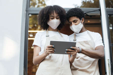 Social media and cafe invitation after lockdown. Young african american female and male in aprons...