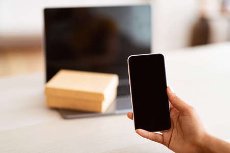 Technology Concept. Unrecognizable black woman holding and showing smartphone with blank empty screen for mock up. Laptop computer with present box or delivery package on keyboard in the backgroundの写真素材