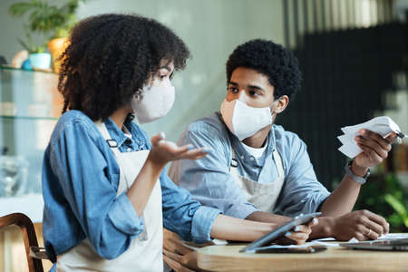 Crisis during covid-19 quarantine, payment of bills and problems in small business. Upset young african american man and woman in protective masks pay taxes, lack of money and gesticulate, free spaceの写真素材